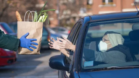 Gloved hands hold out a bag of groceries to a person wearing a mask inside a vehicle